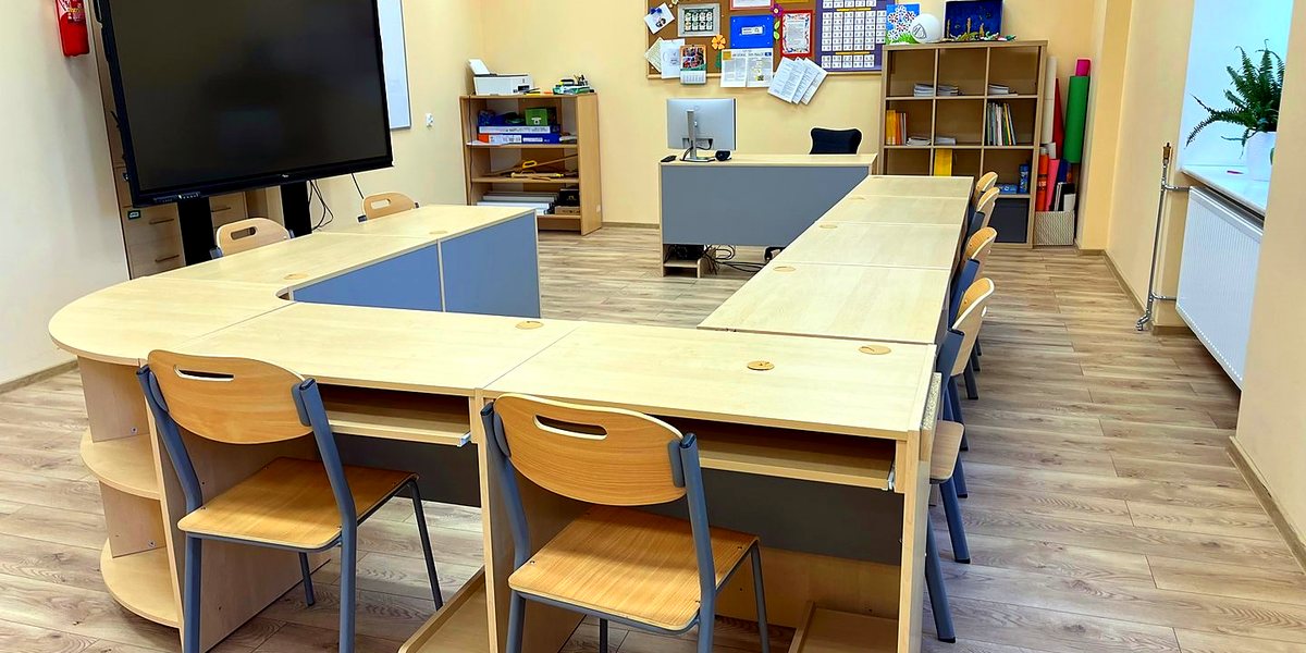 Sala lekcyjna z ławkami ustawionymi w podkowę i monitorem interaktywnym, gotowa do pracy w grupach