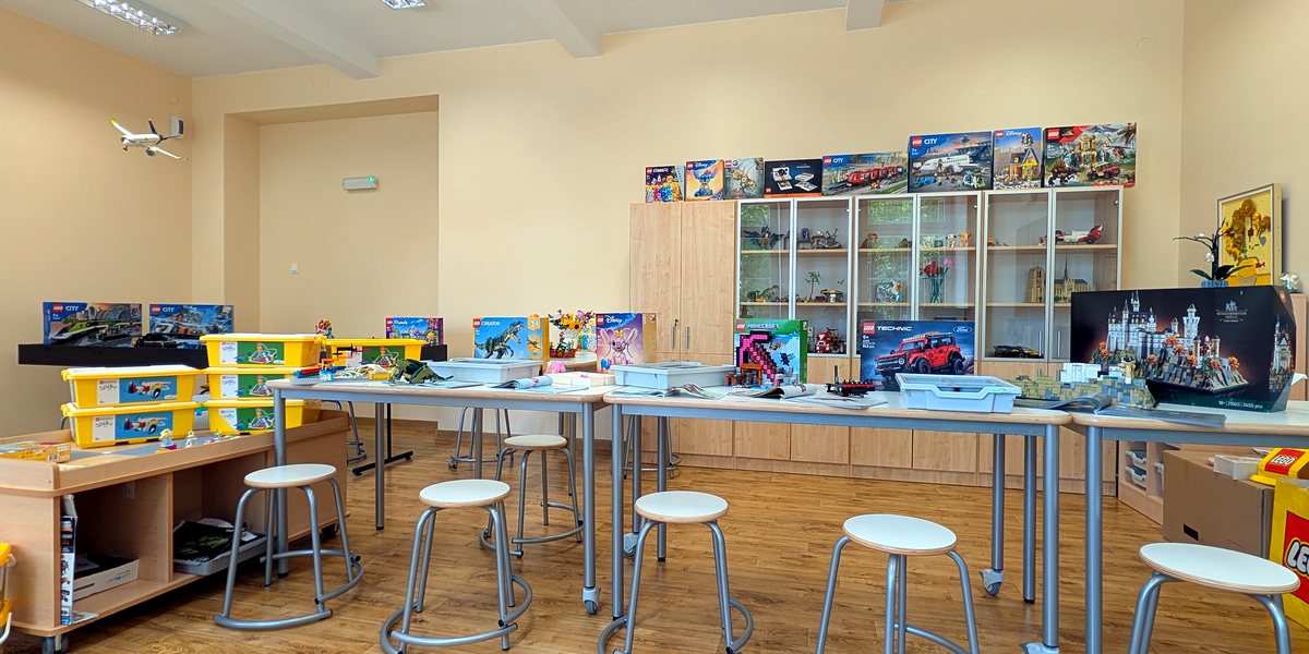 Sala LEGO z długimi stołami do budowania, makietą miasta i regałami pełnymi zestawów klocków